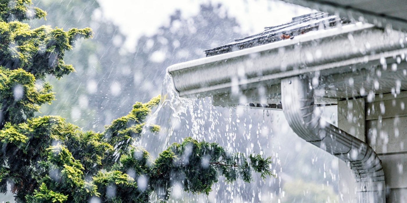 Drenching downpour rain storm water is overflowing off the tile shingle roof - streaming, rushing and splashing out over the overhanging eaves trough aluminum roof gutter system on a suburban residential colonial style house near Rochester, New York State, USA during a torrential mid-summer July downpour.